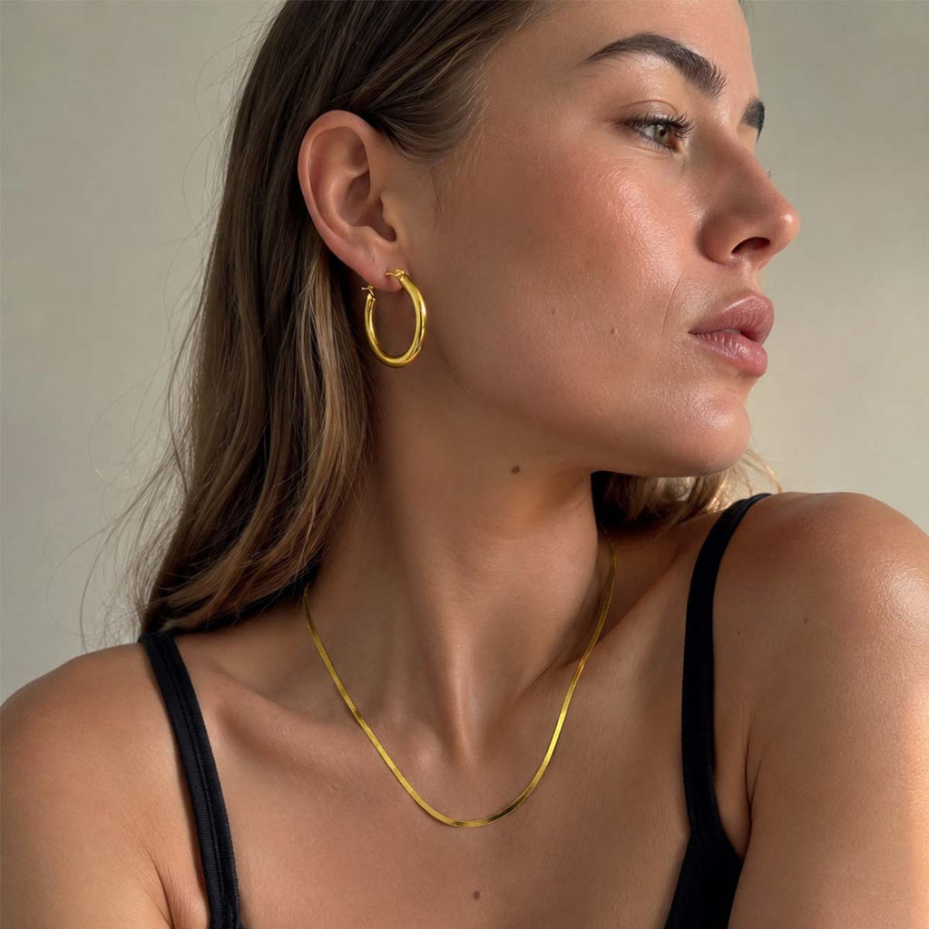 A close-up of a woman wearing a delicate gold chain necklace and a large gold hoop earring, shown from the chest up against a plain beige background.